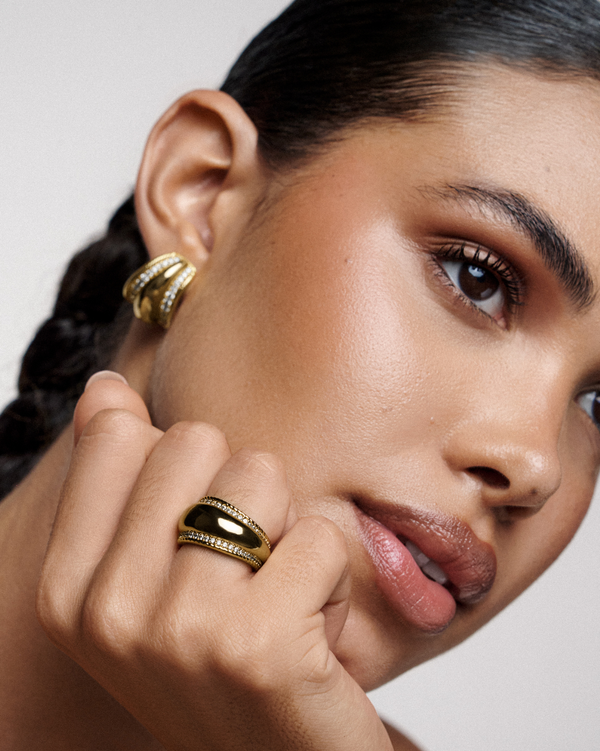 Close-up of a woman wearing gold hoop earrings and a ring, with a neutral background.